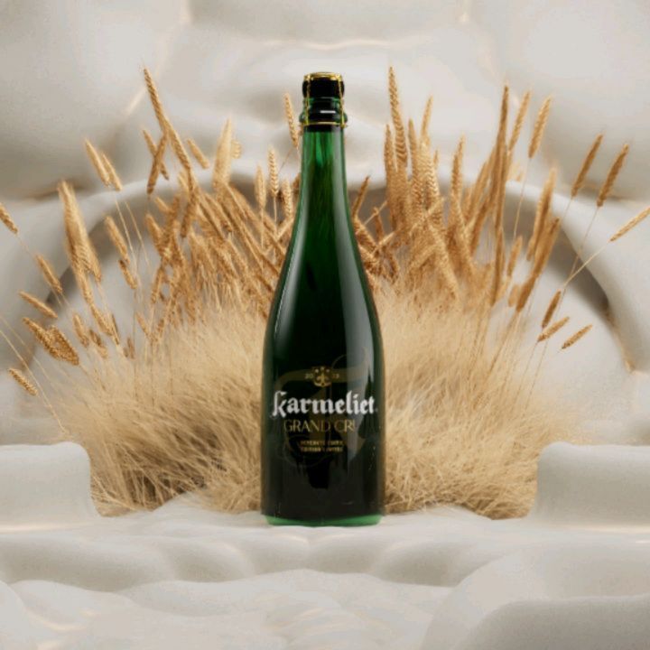 A bottle of Karmeliet Grand Cru beer stands prominently against a textured white backdrop accented by delicate, wheat-colored grasses.