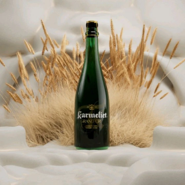 A bottle of Karmeliet Grand Cru beer stands prominently against a textured white backdrop accented by delicate, wheat-colored grasses.