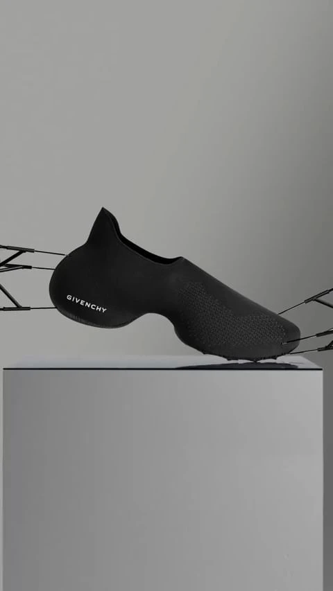 A black Givenchy futuristic slip-on shoe suspended by thin wires above a grey pedestal in a minimalist studio setting.