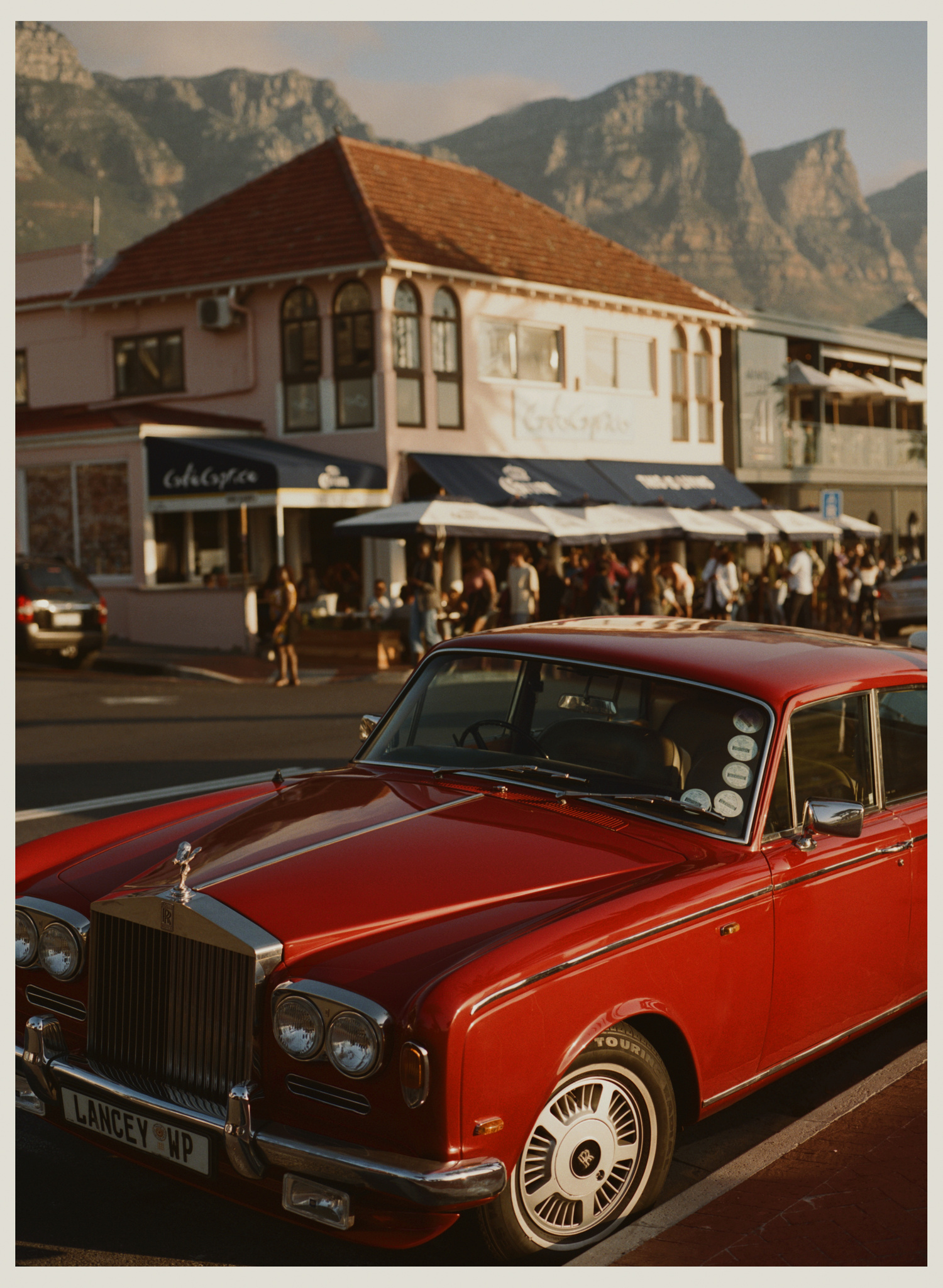 Red Rolls-Royce parked on a city street with a building and mountains in the background during daytime, capturing a vintage and upscale scene.