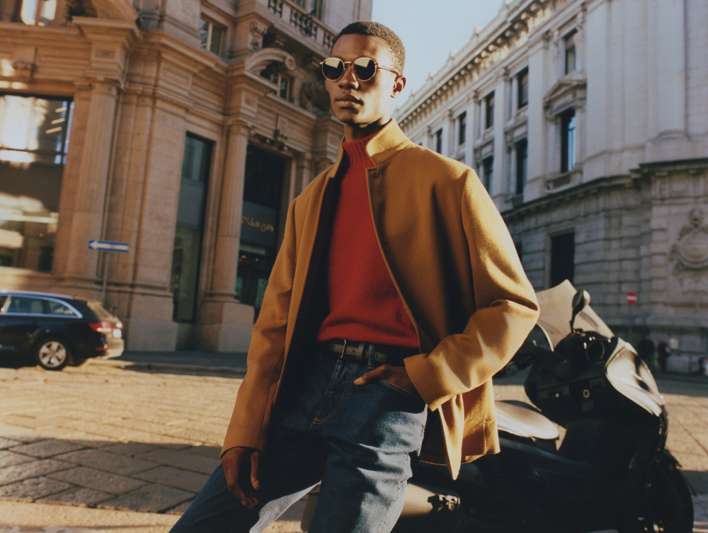 Stylish adult man wearing sunglasses, red turtleneck, tan jacket, and jeans poses with a motorbike on a city street with buildings.