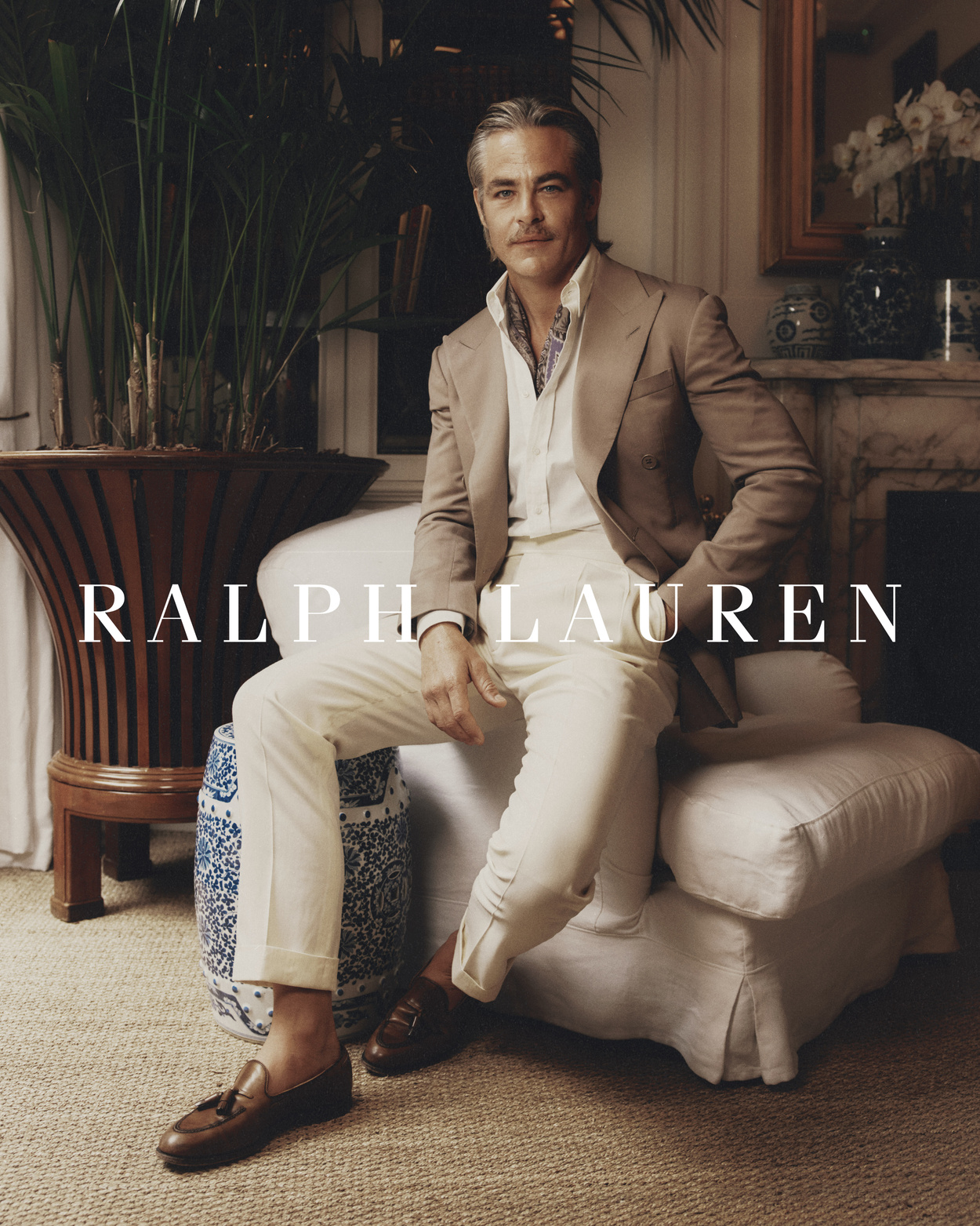 Man in a suit and loafers sits on a cream-colored chair in a room with dark green plants and Ralph Lauren branding. Fashion photography.