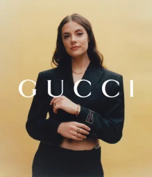 Portrait of a woman wearing a black blazer with Gucci branding, posed against a solid yellow background.