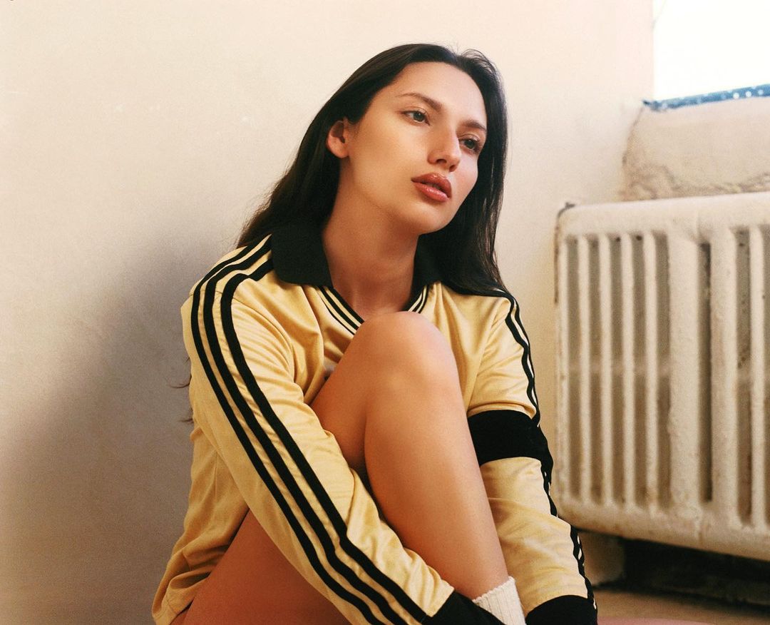 Young woman in yellow athletic long-sleeve shirt sitting on the floor. Radiator visible in the background. Indoor setting, portrait style.