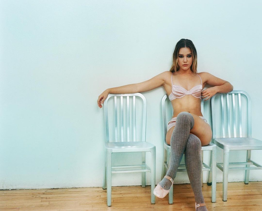 A woman with long brown hair poses in a pink lingerie set and knee-high socks while seated on a metal chair in a studio setting.