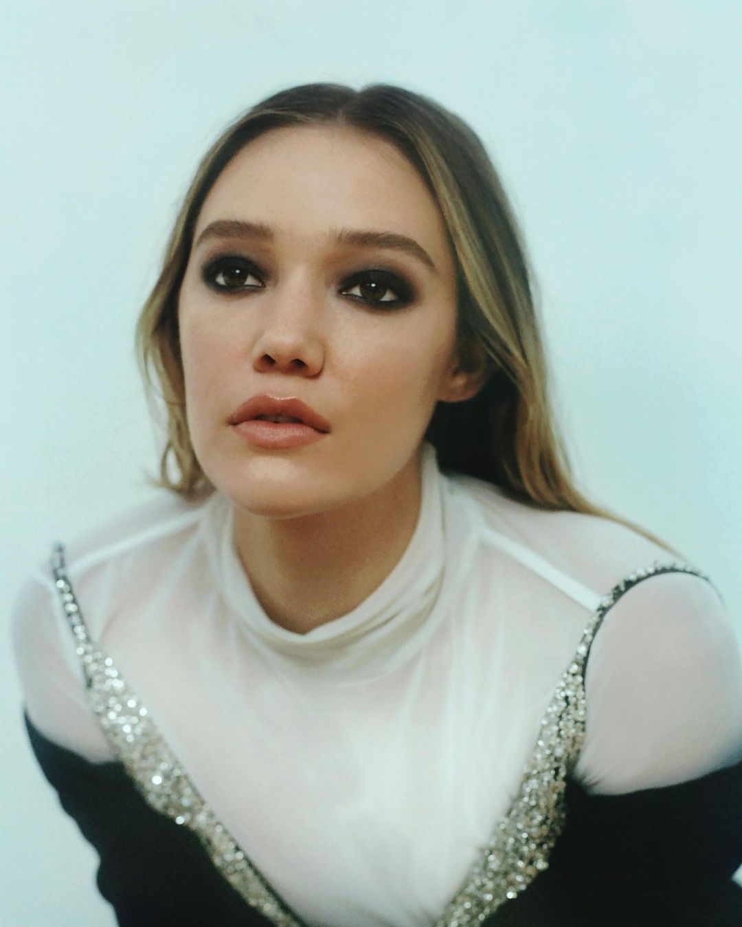 Close-up of a young adult woman with smokey eye makeup, wearing a semi-sheer white top with crystal embellishments, against a light blue background.