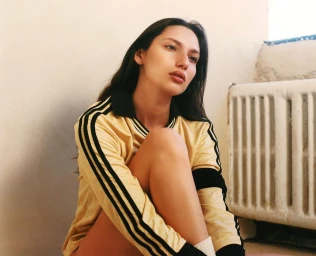 A woman with long dark hair wearing a yellow collared shirt with black stripes sitting near a radiator.