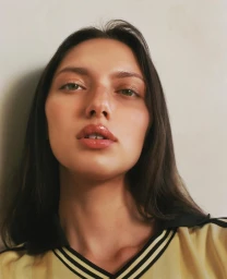 Close-up portrait of a woman with dark hair wearing a yellow collared top against a plain light wall.
