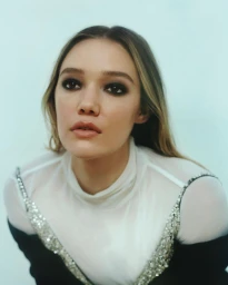 Portrait of a woman with dark smoky eye makeup wearing a white sheer top with embellished straps against a light background.