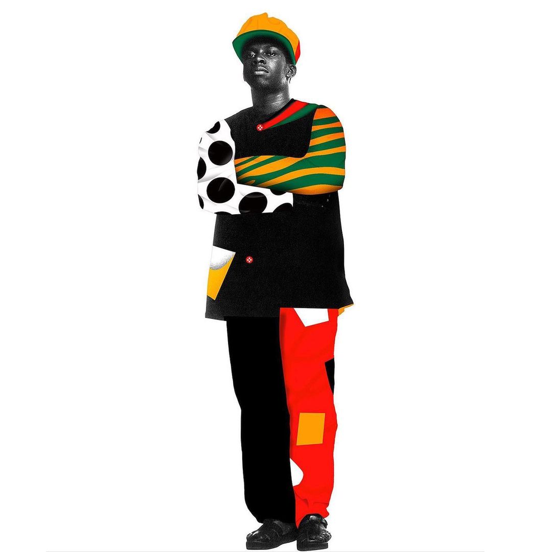 Full-length shot of a stylish black man wearing a yellow hat, patterned jacket, and patchwork trousers against a white backdrop.