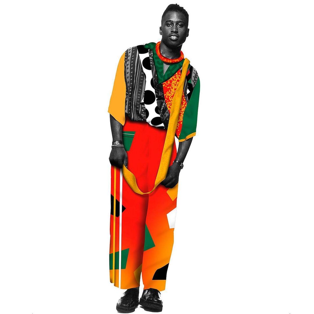 Adult male posing in bright, patterned clothing and black dress shoes against a stark white backdrop in a fashion-forward studio shot.