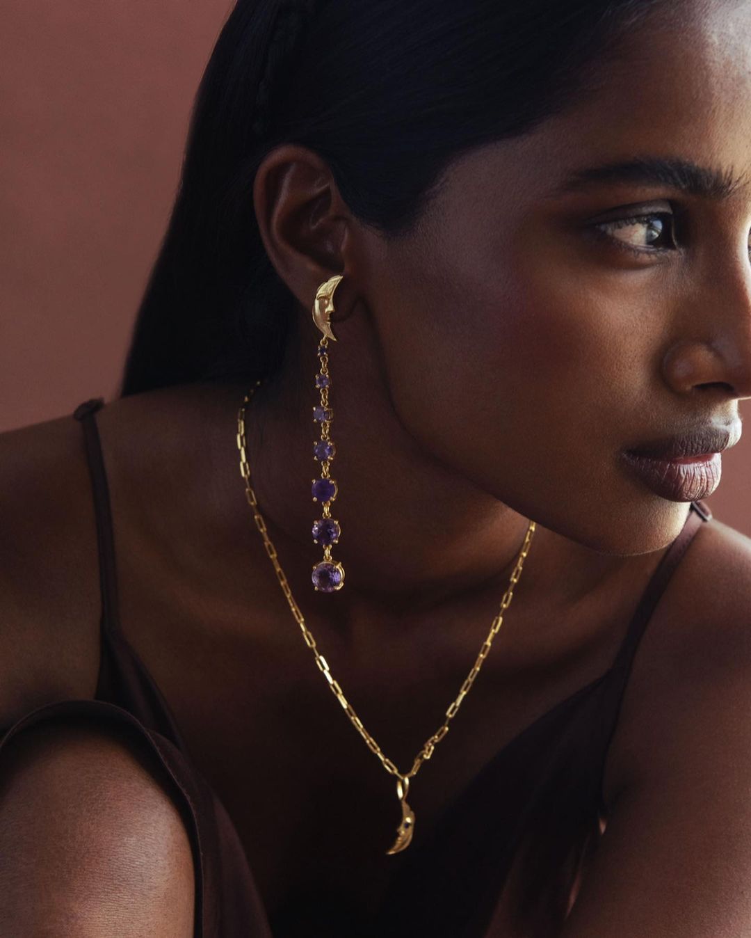 Close-up of an adult female model wearing golden jewelry including earrings with purple stones and a moon pendant necklace.