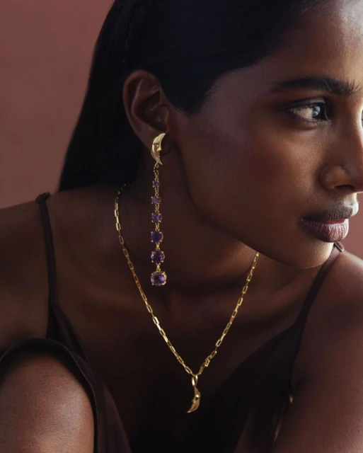 Close-up of an adult female model wearing golden jewelry including earrings with purple stones and a moon pendant necklace.