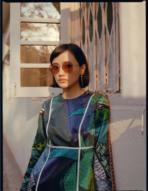 Portrait of an asian woman with round sunglasses wearing a patterned dress. She is standing outside a white building.