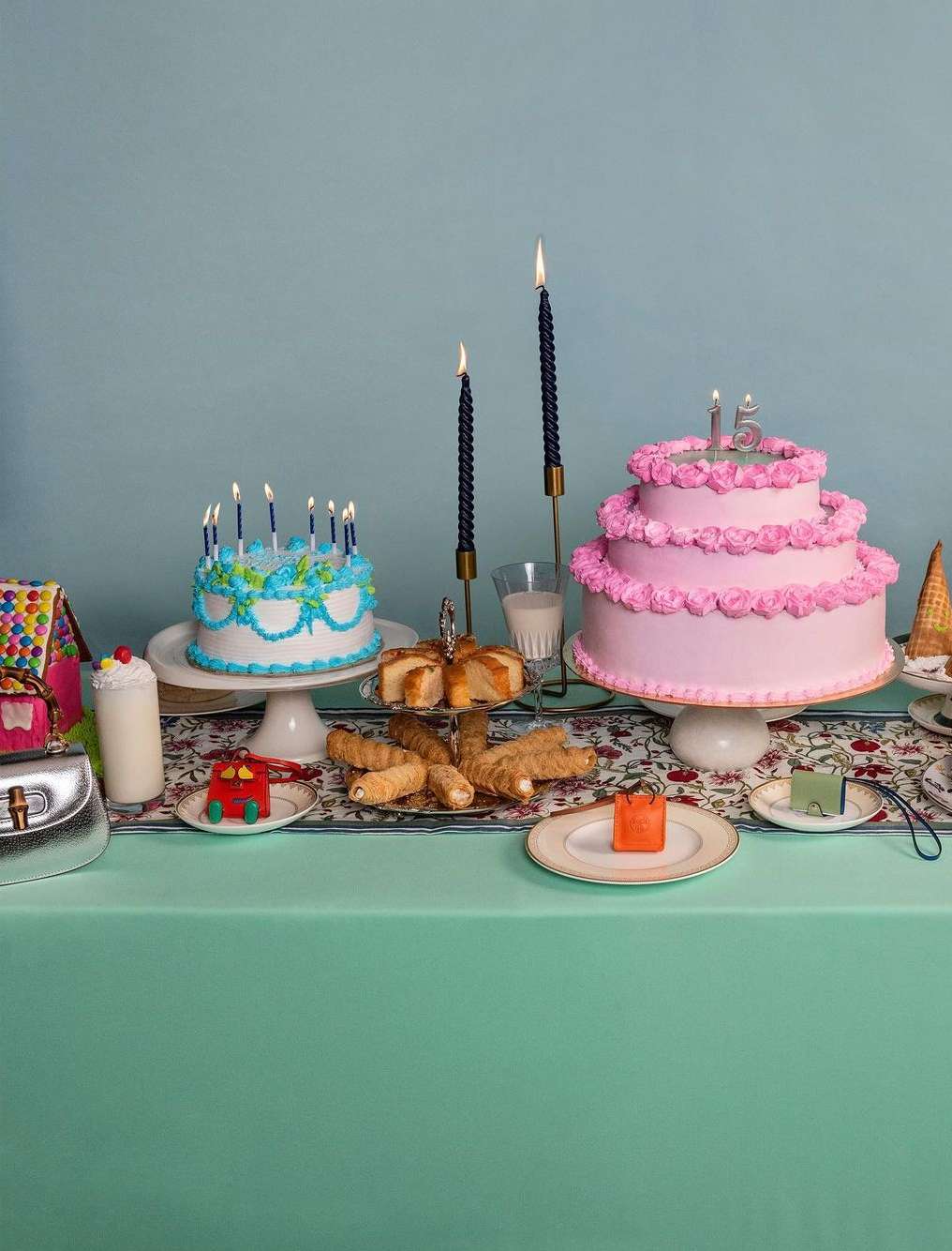 Birthday party scene with pink and blue cakes, candles, and pastries arranged on a floral tablecloth against a teal background.
