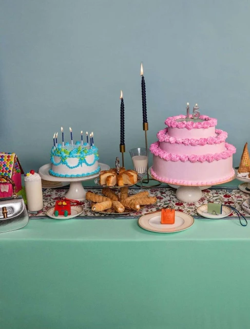 Birthday party scene with pink and blue cakes, candles, and pastries arranged on a floral tablecloth against a teal background.