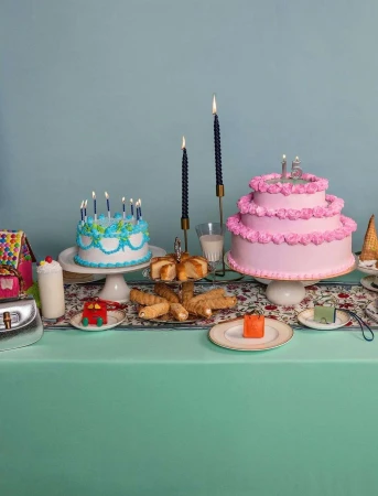 Birthday party scene with pink and blue cakes, candles, and pastries arranged on a floral tablecloth against a teal background.