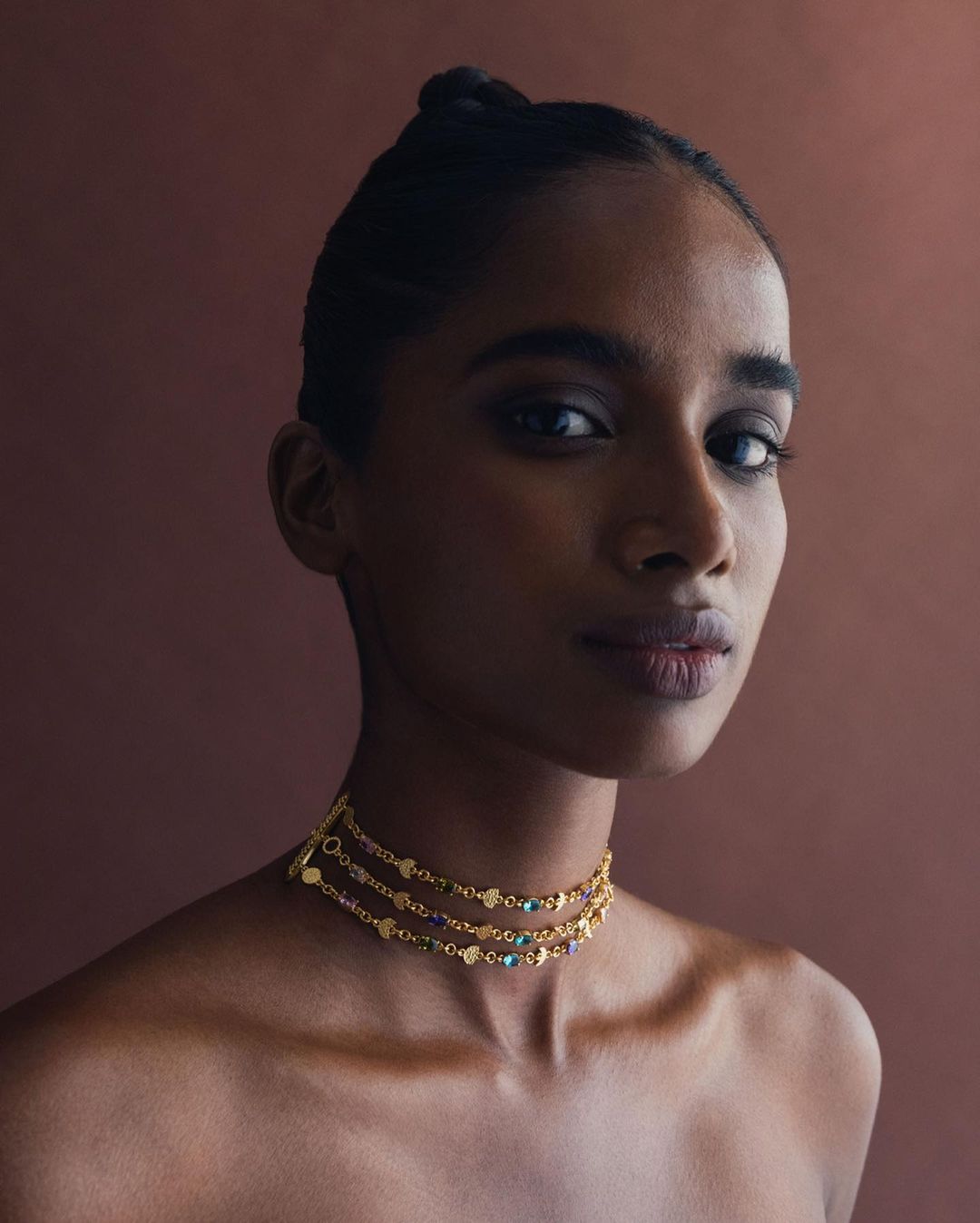 Close-up portrait of a woman with dark hair and a beaded gold choker necklace against a solid brown backdrop in soft studio lighting.