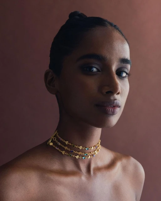 Close-up portrait of a woman with dark hair and a beaded gold choker necklace against a solid brown backdrop in soft studio lighting.