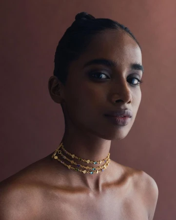 Close-up portrait of a woman with dark hair and a beaded gold choker necklace against a solid brown backdrop in soft studio lighting.