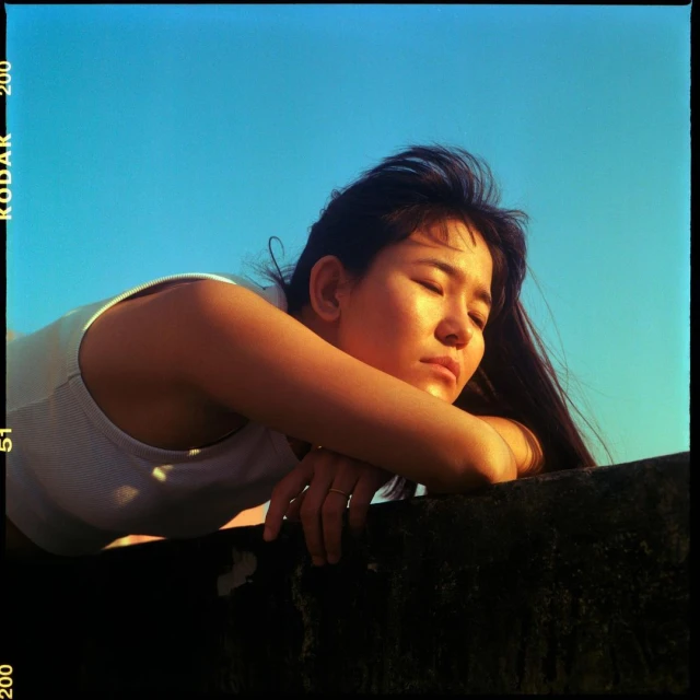 Woman with dark hair rests head on wall under bright blue sky, warm sunlight, and gold ring. Serene outdoor moment in natural light.