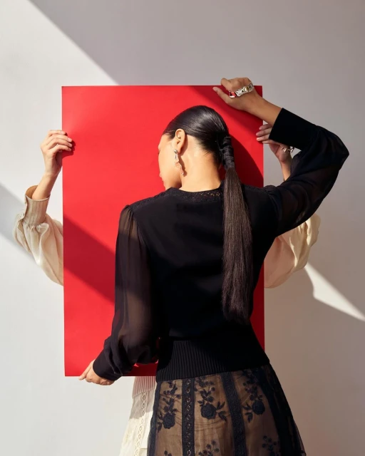 Fashion shot of a woman with a ponytail holding a red board, wearing a black top and skirt, next to another woman in a light-colored top.