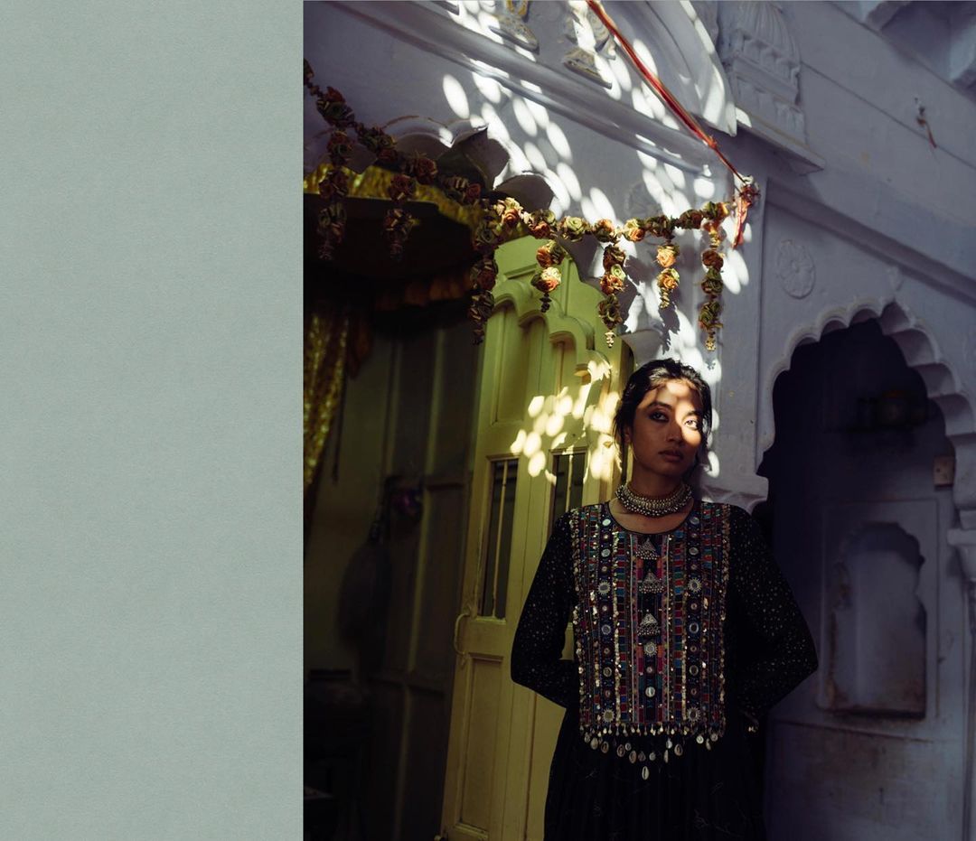 A woman in a black embroidered dress and statement necklace poses in front of a decorated doorway, illuminated by dappled sunlight.