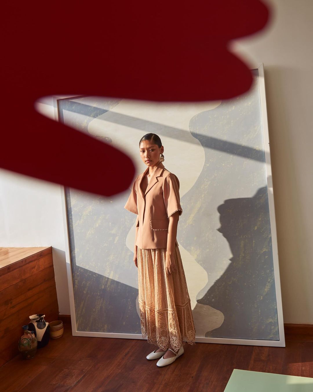 Woman poses indoors wearing tan blazer and lace skirt in front of large abstract painting, accented by shadows and red graphic element.