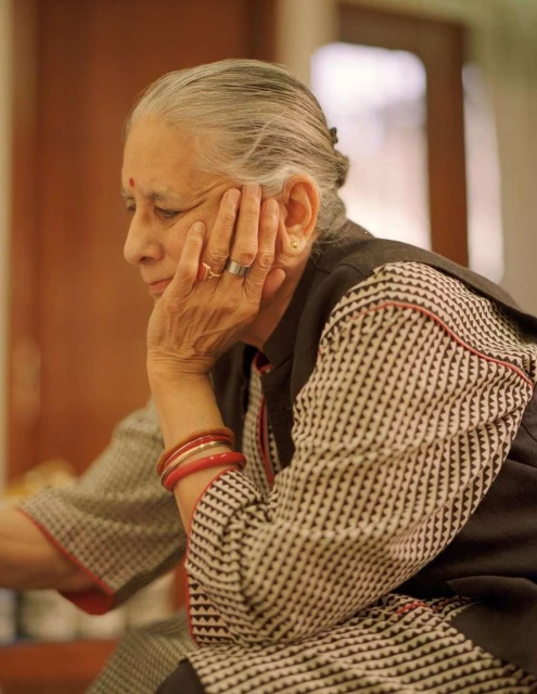 Elderly woman with gray hair rests her face on her hand, wearing a patterned top and bracelets in an indoor setting with neutral tones.