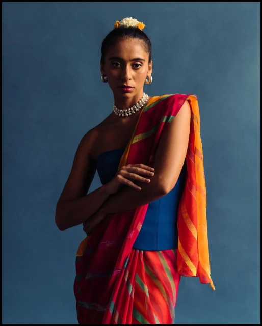 Portrait of a woman with floral hairpiece wearing a blue top and a colorful sari, shot against a deep blue background in studio lighting.
