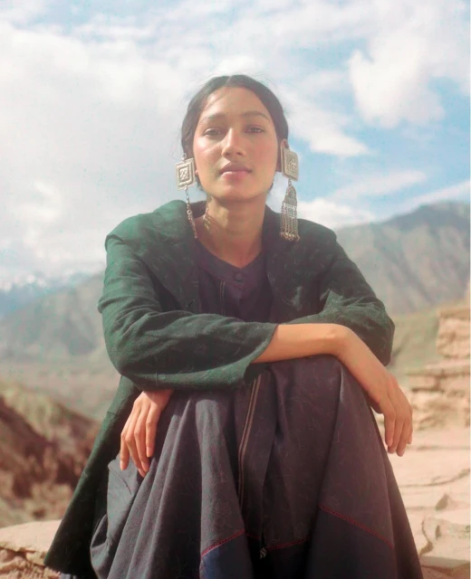 Portrait of a woman wearing traditional jewelry and clothing, seated in front of a mountain range with a cloudy sky background.