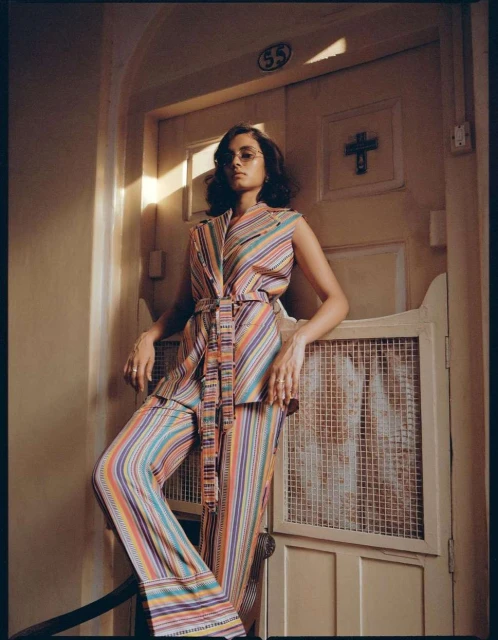 Woman in a striped suit poses on stairs in front of a vintage white door with a cross, bathed in natural light indoors.