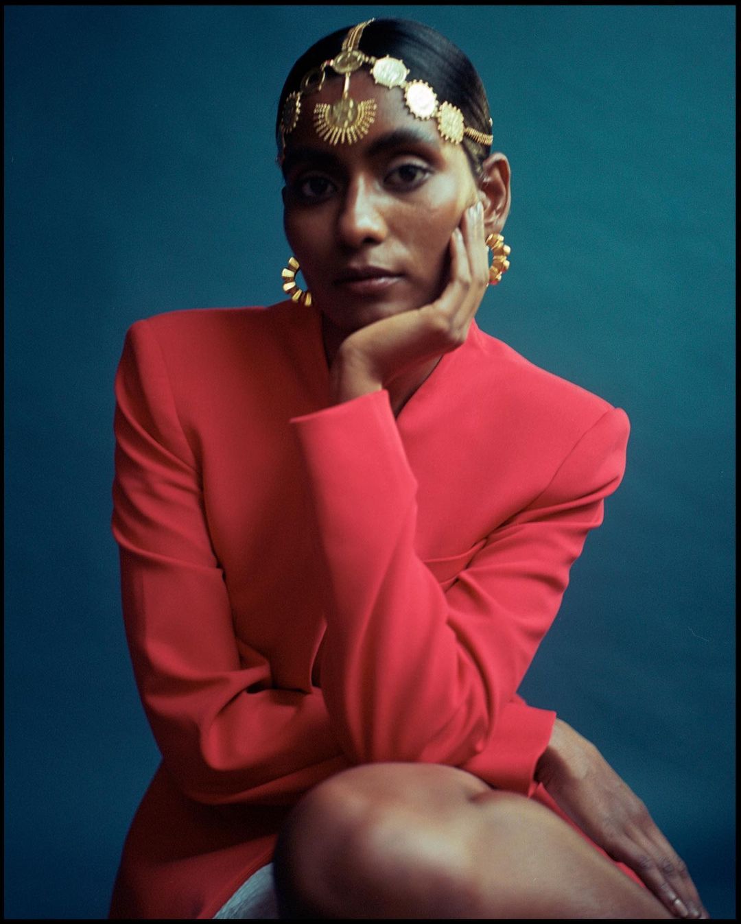 Elegant portrait of a woman with dark hair wearing a red blazer and gold headpiece, set against a teal backdrop in a photo studio.
