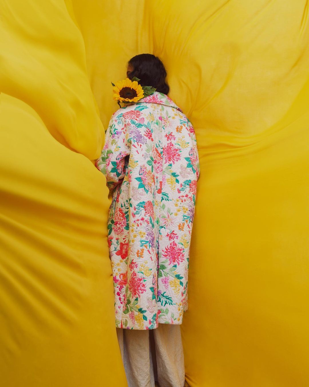 Model in a floral print coat and neutral trousers, carrying a sunflower against a textured yellow backdrop in a fashion editorial.