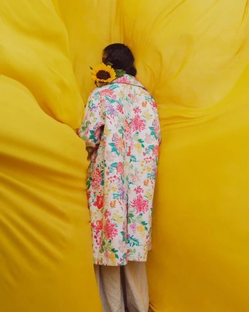 Model in a floral print coat and neutral trousers, carrying a sunflower against a textured yellow backdrop in a fashion editorial.