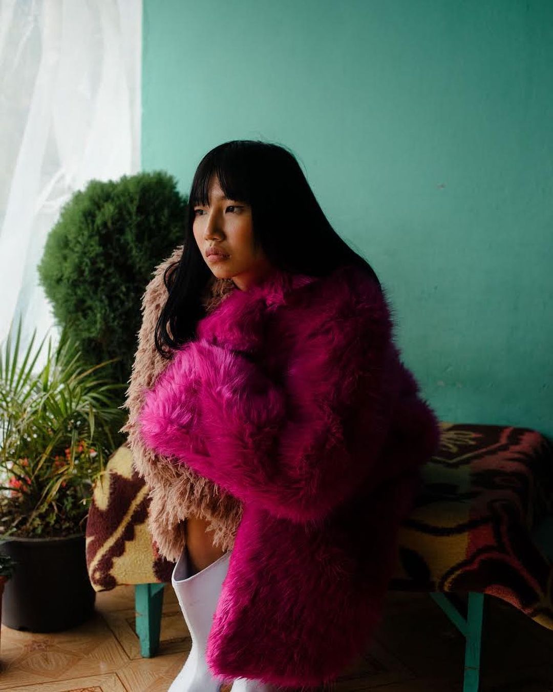 Portrait of an Asian woman with black hair wearing a pink fur coat and white boots, posing indoors near plants and a blanket.