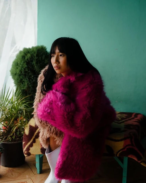 Portrait of an Asian woman with black hair wearing a pink fur coat and white boots, posing indoors near plants and a blanket.
