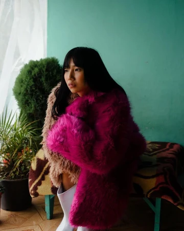 Portrait of an Asian woman with black hair wearing a pink fur coat and white boots, posing indoors near plants and a blanket.