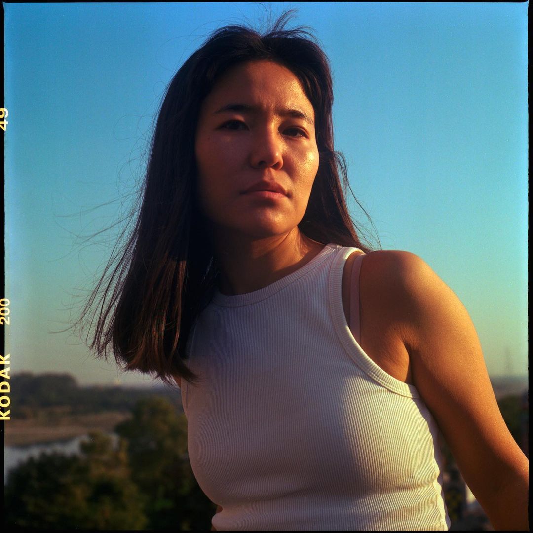 Woman in a white tank top poses outside on a sunny day. The background shows trees, a river, and the bright blue sky.