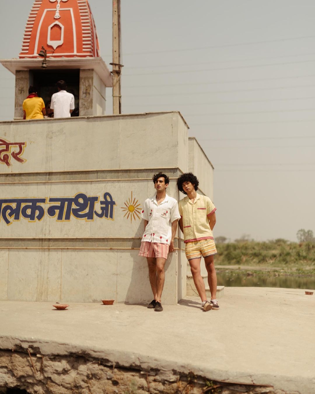 Two young men pose in front of a weathered stone structure with text, wearing casual shirts and shorts in an outdoor setting.