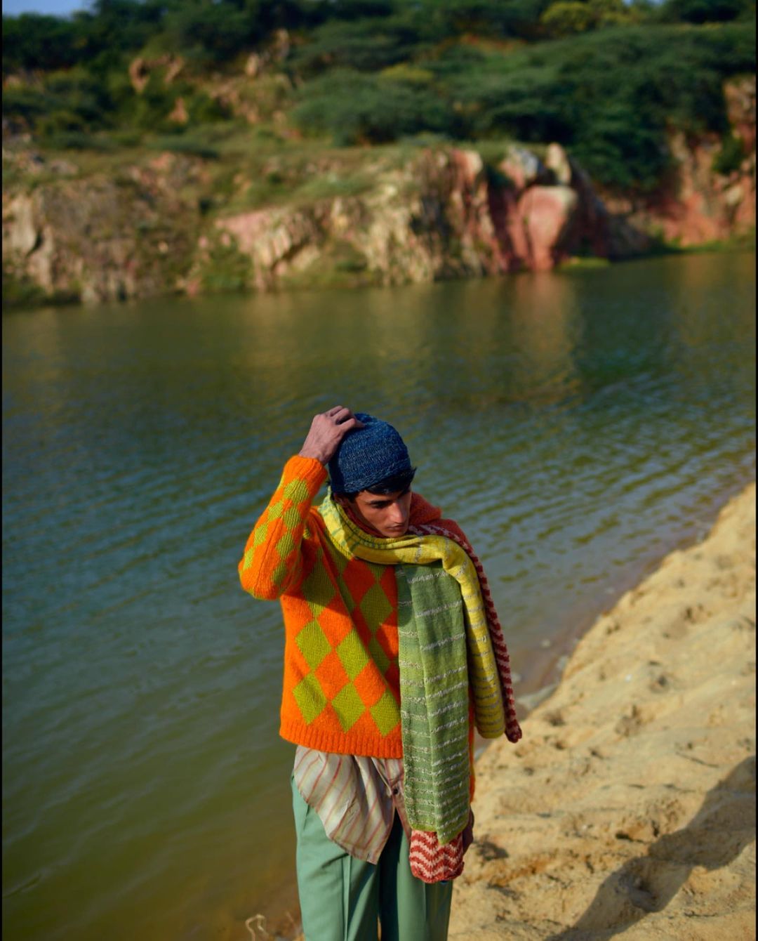 A young man wearing a colorful sweater and hat stands next to a lake, with hills and vegetation visible in the distance.
