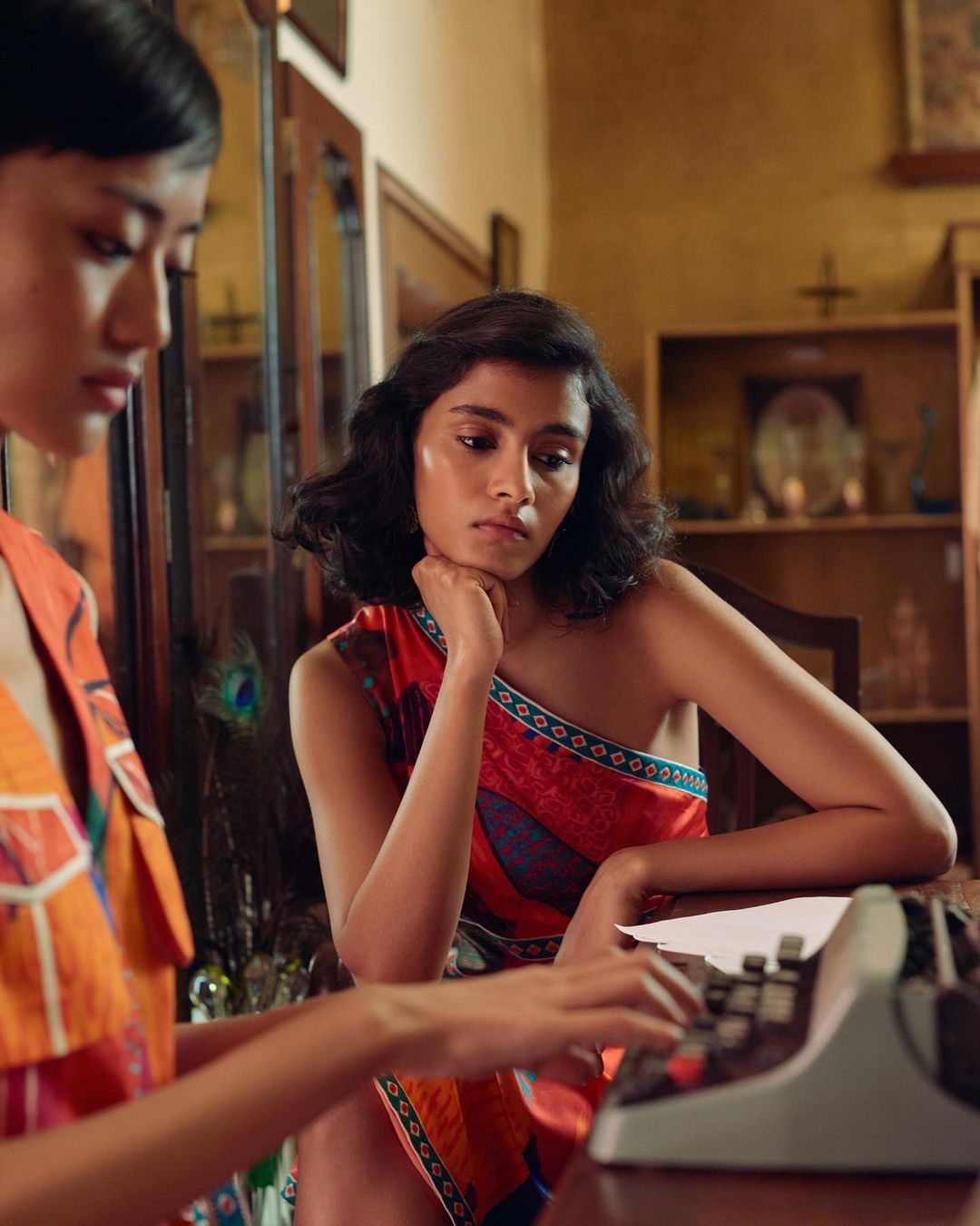 Two women at a desk, one typing on a vintage typewriter, the other resting her chin on her hand in an indoor room setting.