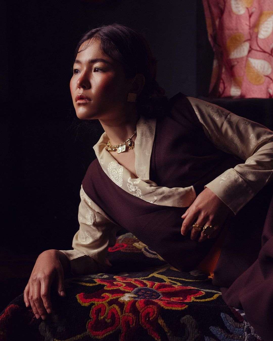 Portrait of a woman in traditional clothing with gold jewelry, sitting indoors with a dark background and patterned fabric visible.