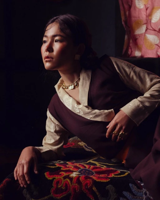 Portrait of a woman in traditional clothing with gold jewelry, sitting indoors with a dark background and patterned fabric visible.