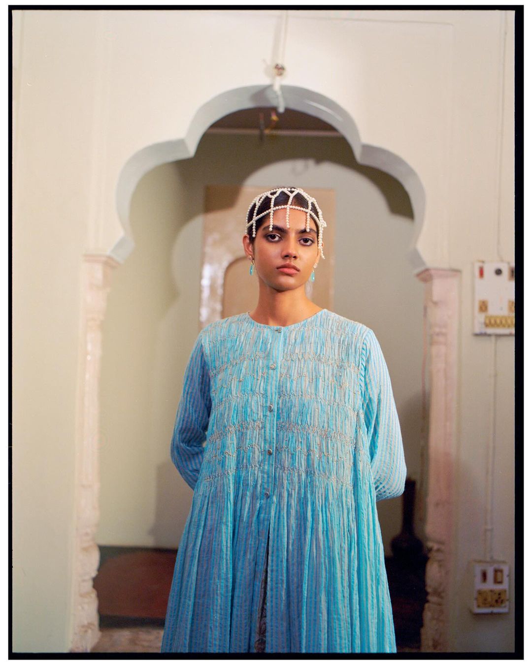 A woman in a blue dress and pearl headpiece stands in an archway, gazing at the camera. The indoor setting features neutral tones.