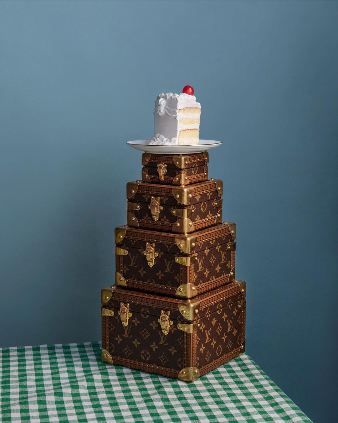 Stacked designer trunks with cake slice. Louis Vuitton branded boxes topped with frosted cake and cherry against a blue background.