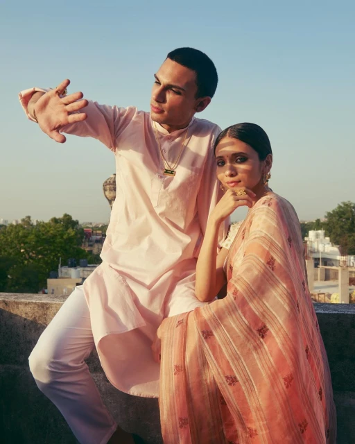 Two people wearing traditional South Asian clothing pose on a rooftop with city views in the background in soft, natural light.