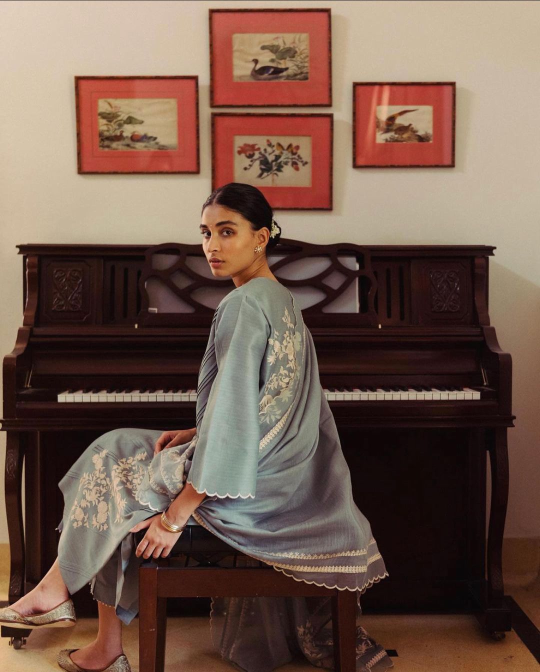 Adult woman sits on a piano bench wearing an embroidered tunic top and wide-leg pants in front of a piano with framed artwork on the wall.