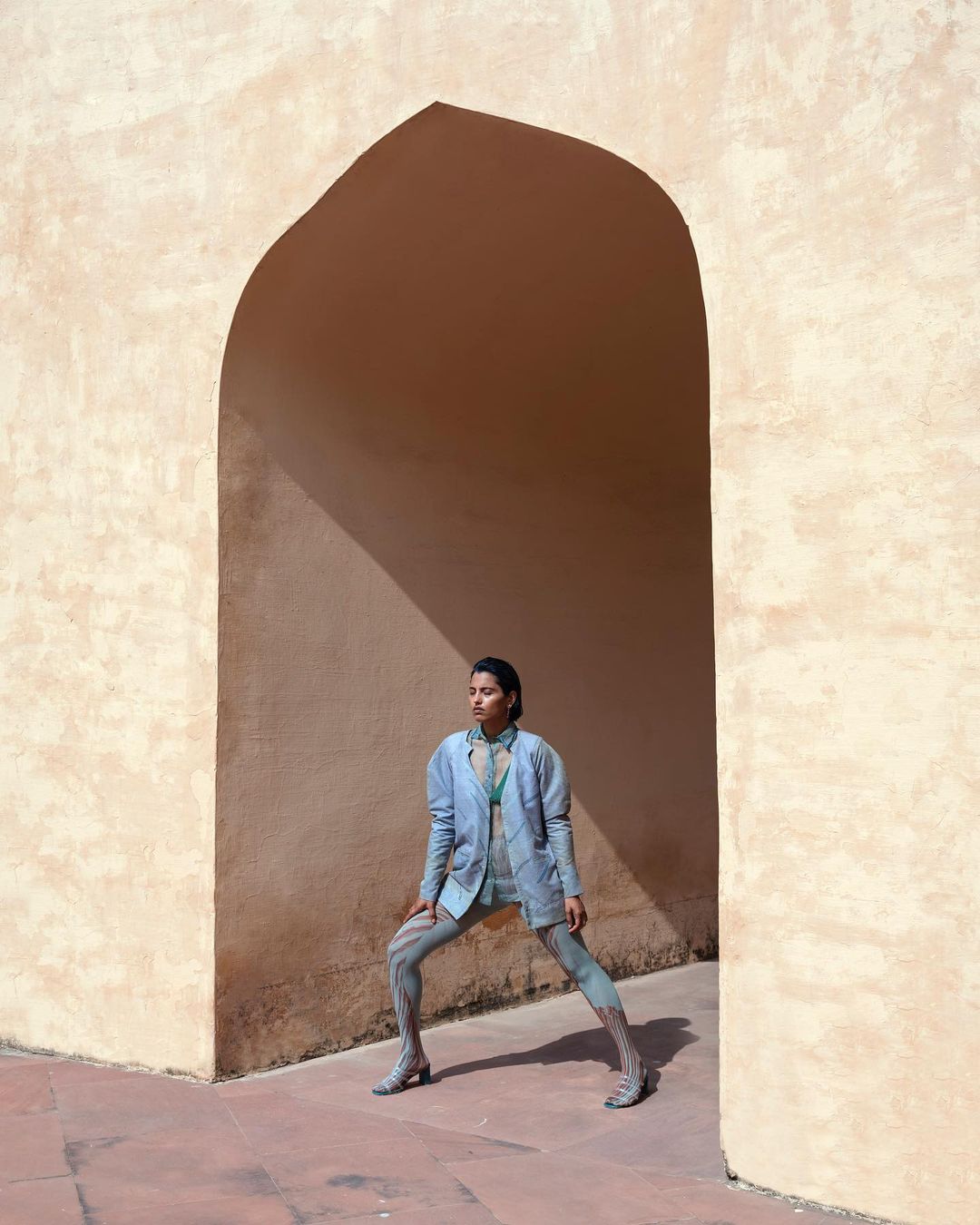 A stylish adult female model in a blue blazer and leggings poses in a desert archway with a serious expression and natural lighting.