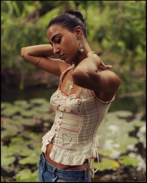 Woman in tank top poses near lily pad pond. Medium shot. Focus on fashion and nature.